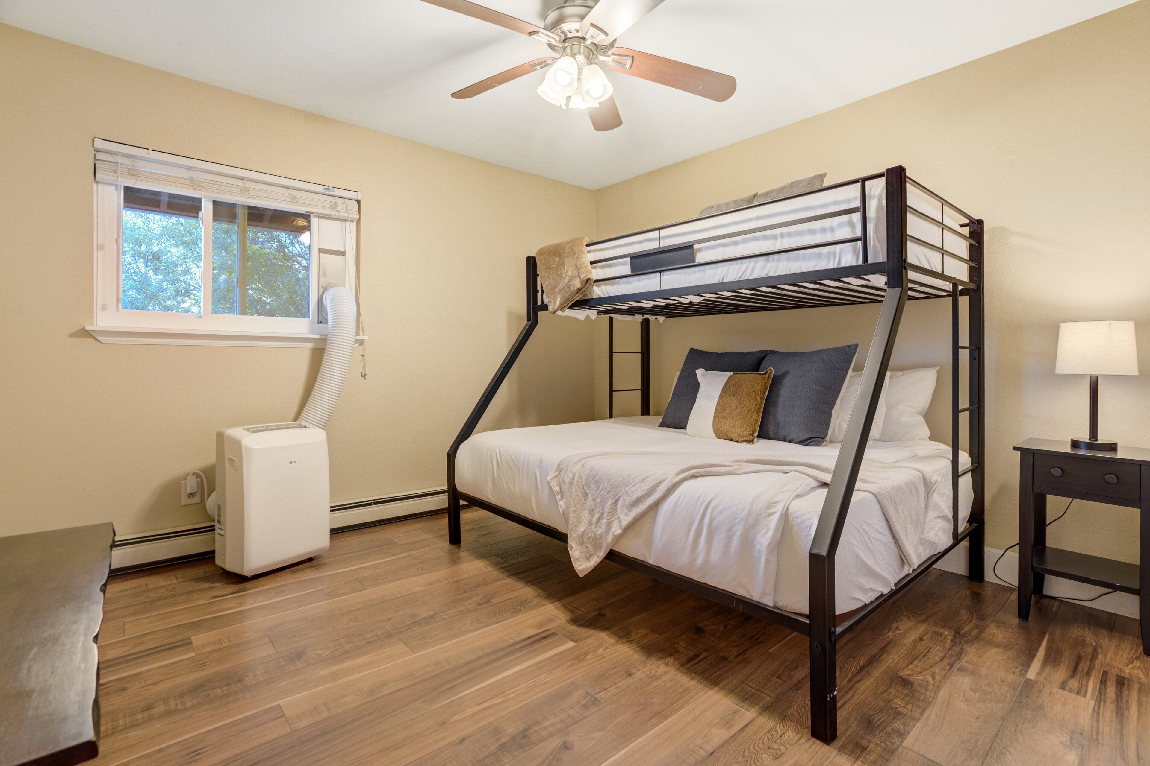 A bedroom with a bunk bed, a ceiling fan, a heater, a window with blinds, and a bedside table with a lamp.