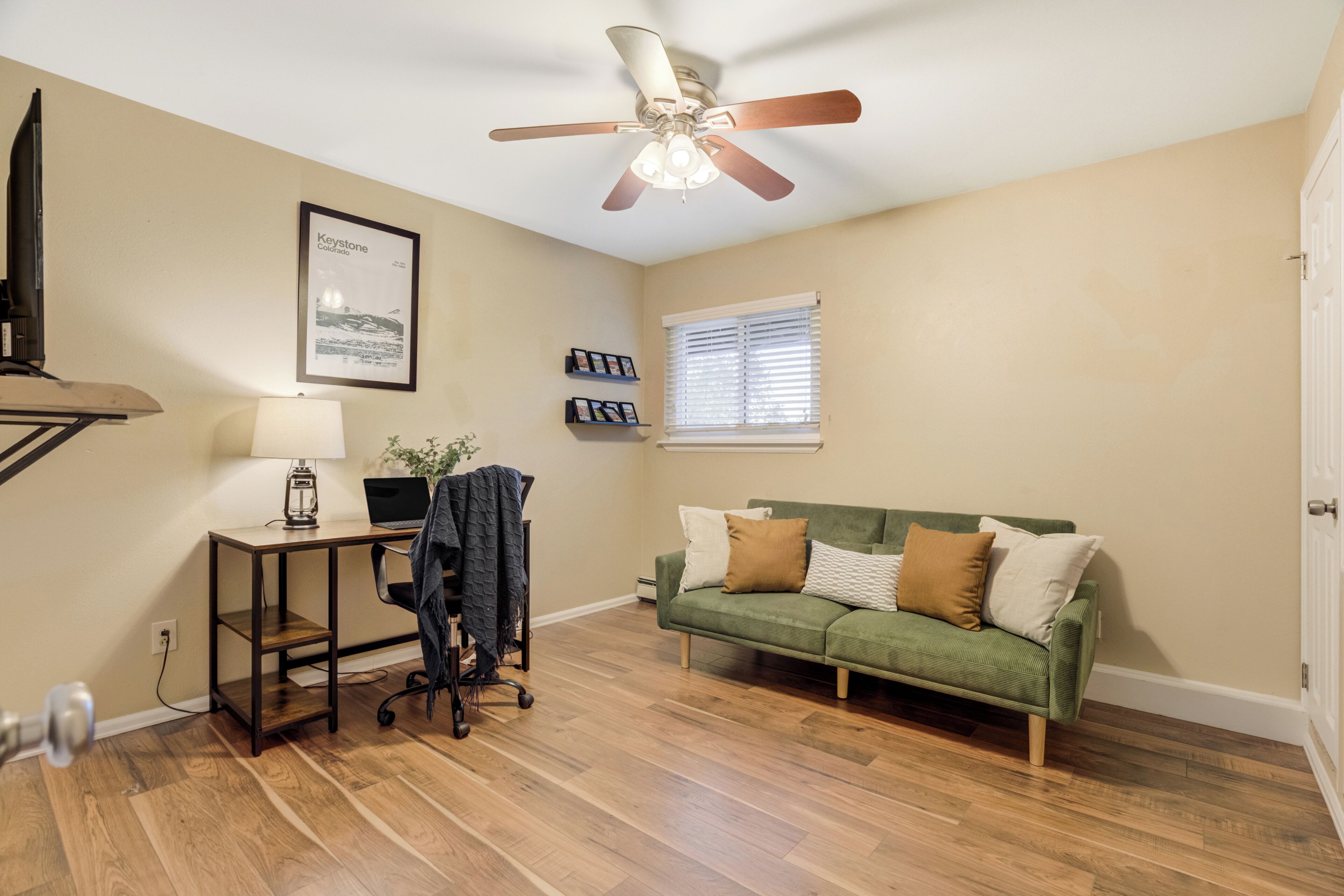 A room with a ceiling fan, a desk with a lamp, a laptop, a green sofa, and a framed picture on the wall.