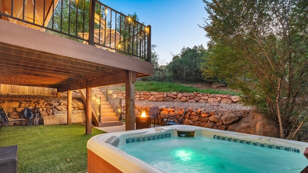 A hot tub with a wooden deck and stone wall backdrop, a staircase leading to an upper deck, and outdoor seating. - 3754 Red Canon (Colorado Springs)