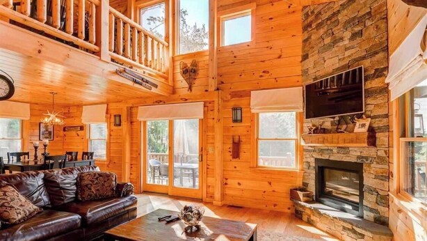 A spacious cabin interior with a stone fireplace, wooden walls, and a balcony. - 489oer (Whitefield)