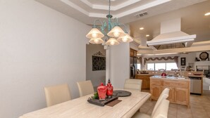 Un coin repas avec une longue table, plusieurs chaises et un lustre. On aperçoit une cuisine à l’arrière-plan, équipée d’appareils électroménagers modernes et dotée d’une fenêtre.
