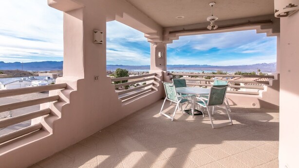 Un balcon avec une table et des chaises, une vue sur les montagnes et un plan d’eau, ainsi qu’un ventilateur de plafond.