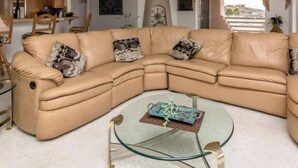 Un canapé d’angle en cuir marron avec des coussins à motifs, une table basse en verre agrémentée d’un objet décoratif, et une vue sur un paysage montagneux par une grande fenêtre.