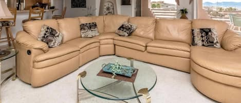 A tan leather sectional sofa with patterned cushions, a glass coffee table with a decorative item, and a view of a mountainous landscape through a large window.