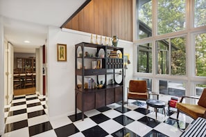 A room with a checkered floor, a wooden bookshelf, two chairs, and a large window with a view of trees. - Pineview Haven (Knoxville)
