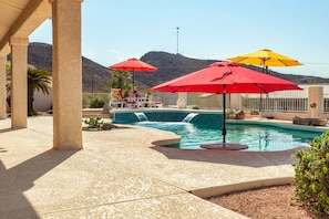 Zona de piscina con sombrillas rojas y amarillas, piscina con baldosas y montañas al fondo.