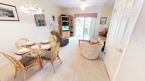 A dining area with a glass table set for four, surrounded by wicker chairs. A television is mounted on a wooden shelf, and a sofa is visible in the background. - Village at the Glens G 109 (Little River)