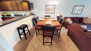 A dining area with a wooden table set for four, surrounded by chairs. A kitchen area with wooden cabinets and a countertop is visible to the left. - Village at the Glens S 205 (Little River)