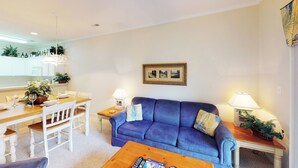 A living room with a blue sofa, a wooden coffee table, a dining table with chairs, and a kitchen in the background. - Village at the Glens K 102 (Little River)