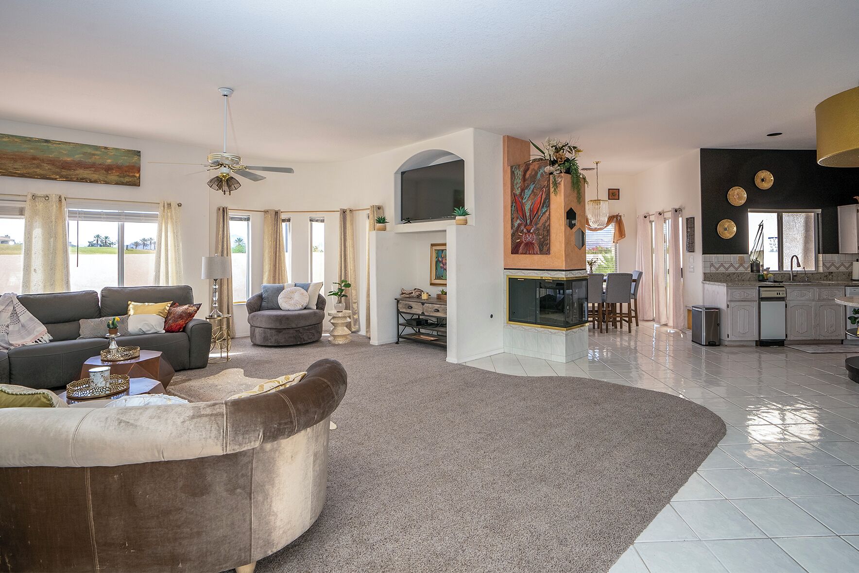 A spacious living area with a grey sofa, a brown armchair, and a grey armchair with a white cushion. There is a flat-screen TV mounted on the wall, a dining area with a table and chairs, and a kitchen with a sink and stove.