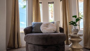 Un sillón mullido de color oscuro con un otoman redondo, dos almohadas y una planta decorativa en el alféizar de una ventana.
