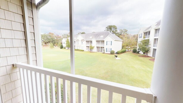 A view from a balcony overlooking a well-maintained lawn with a golf cart, a large white building with multiple windows and balconies, and a clear sky. - Village at the Glens C 204 (Little River)