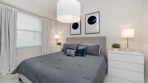 Een slaapkamer met een groot bed, witte nachtkastjes, een grijze hoofdeinde en twee wandlampen.