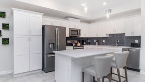 Een moderne keuken met witte kastjes, roestvrijstalen apparatuur, een centraal eiland met barkrukken en een zwart betegelde spatwand.