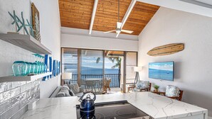 A modern kitchen with a large island, a ceiling fan, and a view of the ocean through large windows. - Maalaea Banyans 411 (Wailuku)