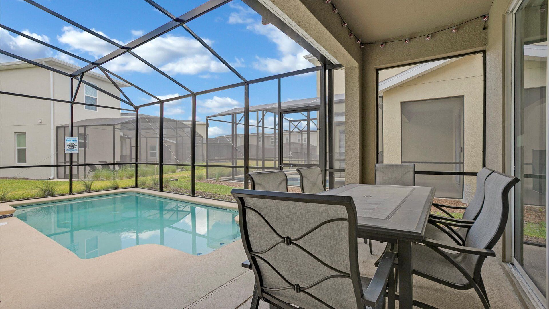 Un espace aménagé au bord de la piscine, avec une table et des chaises, offrant une vue dégagée sur la piscine, ainsi qu’une terrasse couverte donnant sur les bâtiments voisins.