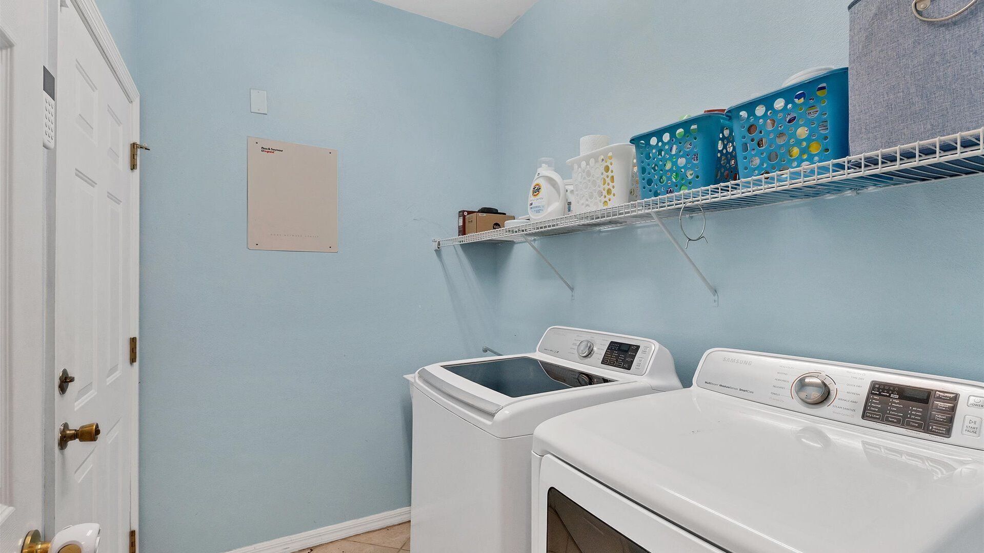 Una lavandería con dos lavadoras y secadoras de carga frontal blancas, una pared azul, una puerta blanca y un estante con productos de lavandería.