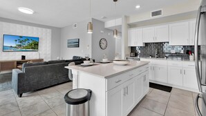 A modern kitchen with white cabinets, a stainless steel refrigerator, and a dining area with a gray sofa and a flat-screen TV. - Pxc3151-306 (Kissimmee)