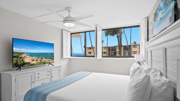 Een slaapkamer met een groot bed, een plafondventilator, een televisie aan de muur en een raam met uitzicht op palmbomen en gebouwen.