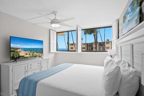 Un dormitorio con una cama grande, un ventilador de techo, un televisor en la pared y una ventana con vistas a palmeras y edificios.