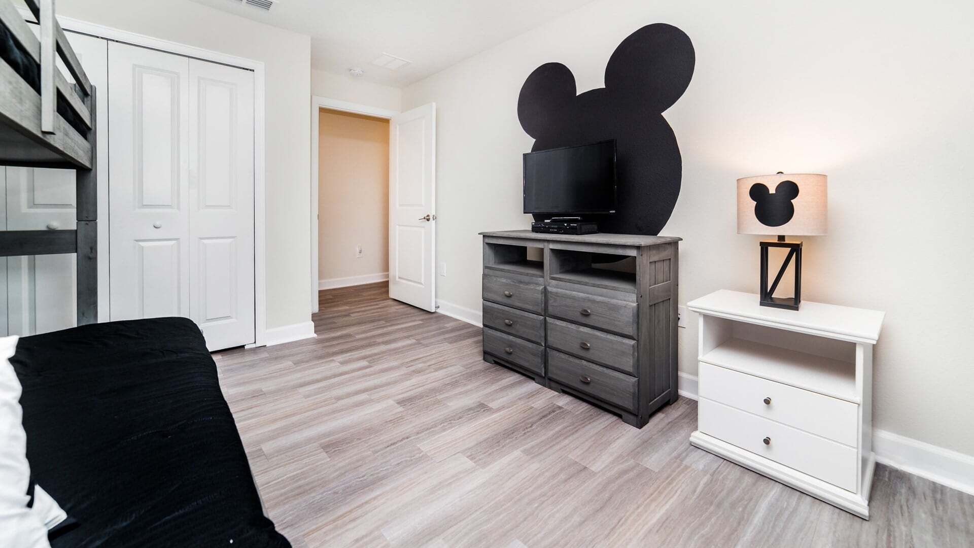 A bedroom with a bed, a dresser, a TV on top, a Mickey Mouse cutout on the wall, and a nightstand with a lamp.
