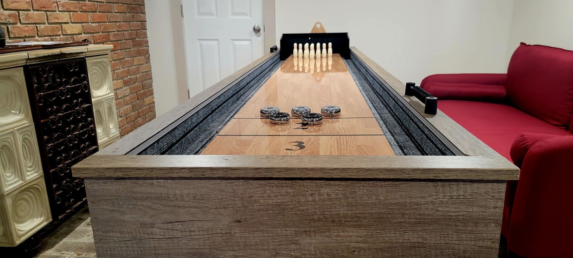 A shuffleboard table with a number 3 marker, a red couch, and a brick wall.