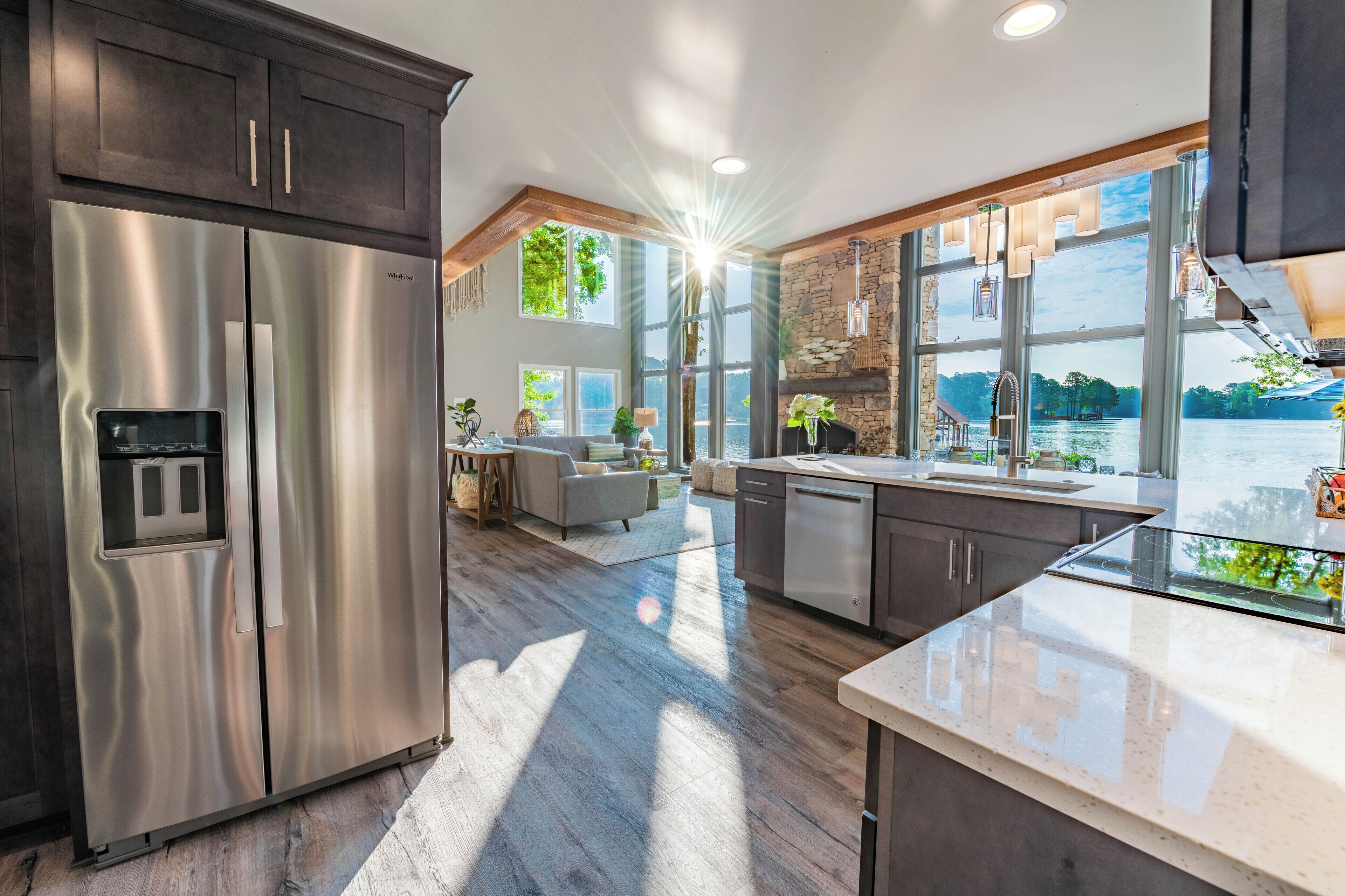 A modern kitchen with a stainless steel refrigerator, wooden flooring, and large windows overlooking a lake.