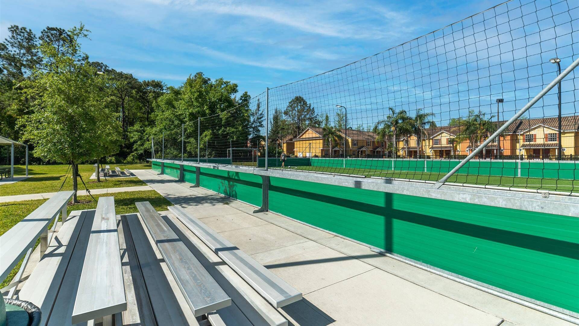 A tennis court with a green fence, benches, and houses in the background.