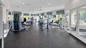 Ein Fitnessraum mit verschiedenen Trainingsgeräten, großen Fenstern und einem Deckenventilator.