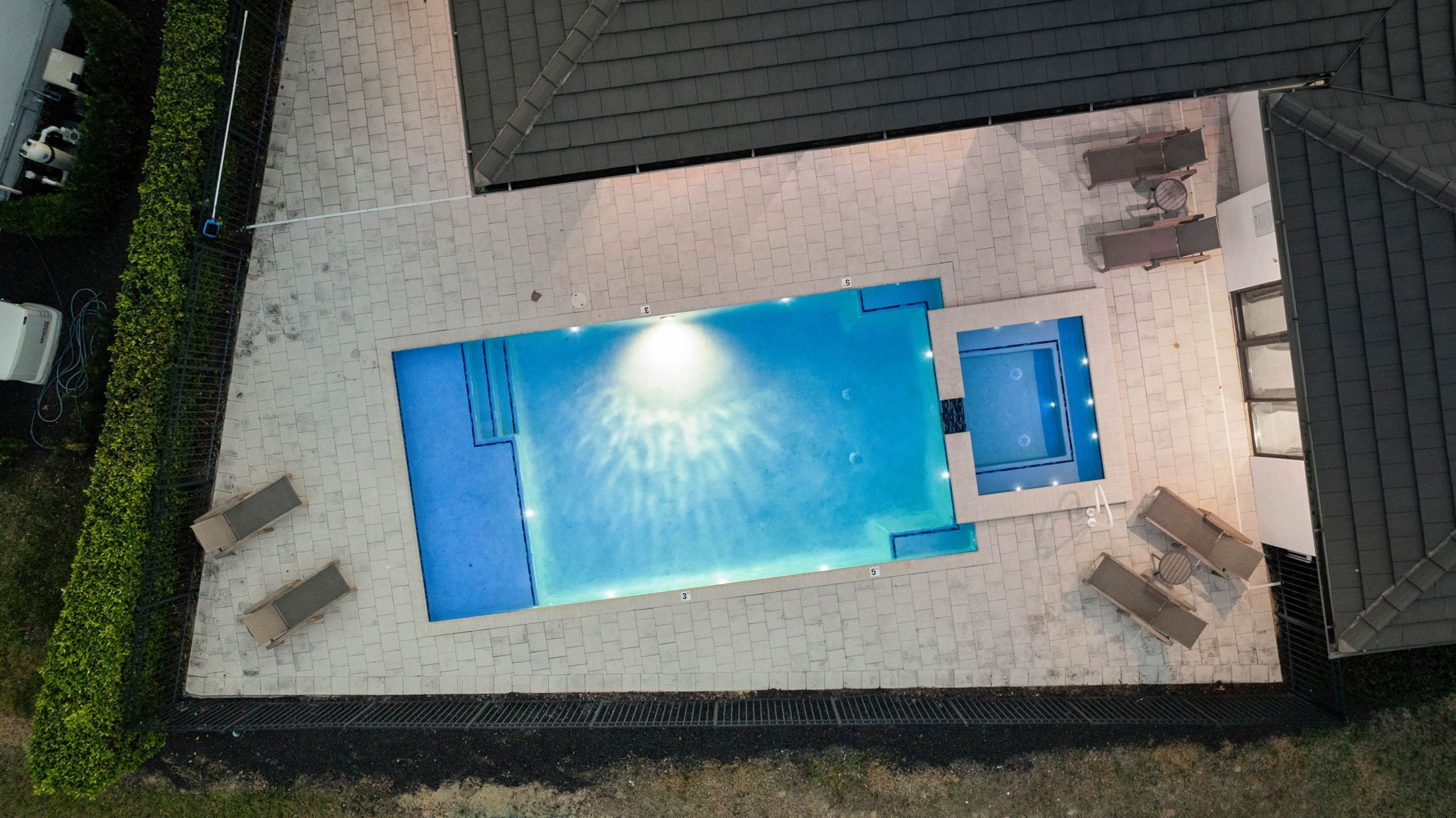 An aerial view of a rectangular swimming pool with a sunken center, surrounded by a paved deck and a low brick wall.
