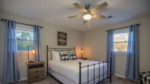 A bedroom with a bed, a nightstand, a lamp, a ceiling fan, and two windows with curtains. - 941 Pine Manor Family (Marietta)