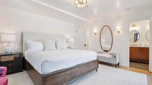 Une chambre moderne avec un grand lit, des tables de chevet, une commode et une salle de bain équipée d’un miroir et d’un lavabo.