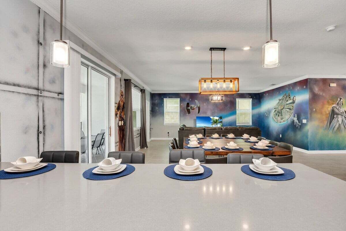 Un comedor moderno con una larga mesa preparada para una comida, un mural con motivos espaciales en la pared y un rincón de estar informal al fondo.