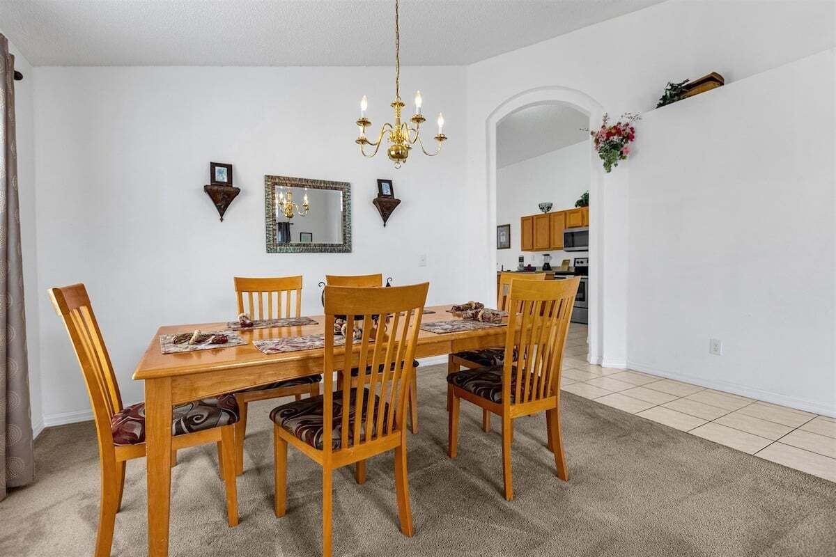 A dining area with a wooden table and chairs, a chandelier, and a kitchen in the background.