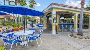 Een terras met blauwe en witte stoelen, een grote blauwe parasol, een overdekte bar en een klok aan de muur.