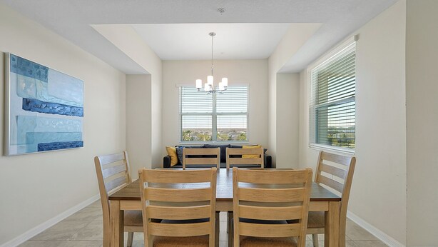 A dining area with a wooden table and chairs, a chandelier, and a large window with blinds. - 3161 Paradox Circle, Unit 406 Kissimmee, 2 Beds (Kissimmee)
