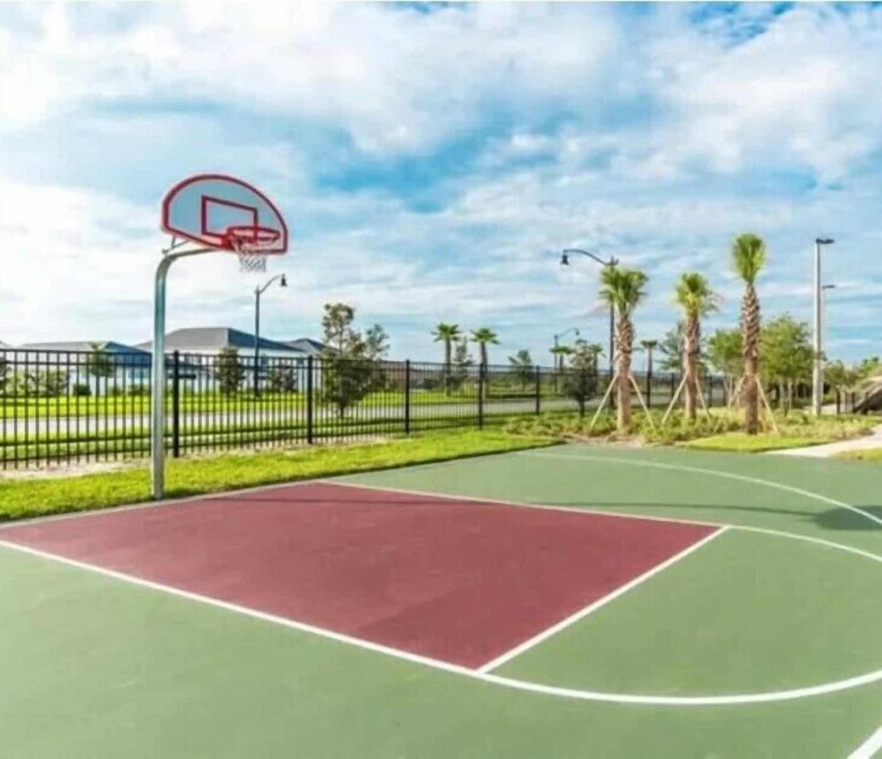 An outdoor basketball court with a red and green surface, surrounded by a black fence, palm trees, and a clear sky.
