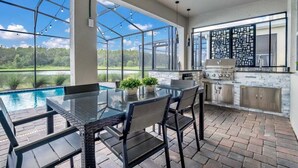 An outdoor kitchen with a glass table and chairs, a barbecue grill, and a view of a lake. - 3017ps · Brand New Luxury 9BR Villa/ Pool/spa/game Room (Kissimmee)