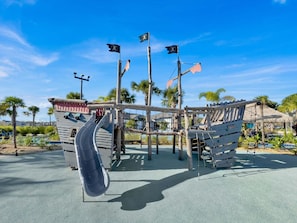 Un parque infantil con tobogán y estructuras para escalar, rodeado de palmeras y un cielo despejado.
