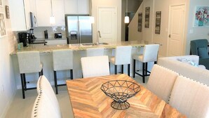 A modern kitchen with white cabinets, a stainless steel refrigerator, and a dining area with a wooden herringbone table and white upholstered chairs. - Special Offer Townhome at Compass Bay (Kissimmee)