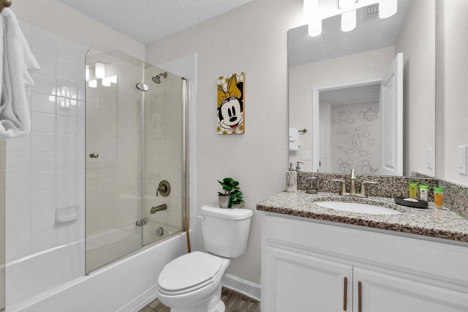 Un baño con ducha envuelta en cristal, inodoro, lavabo con encimera de granito y espejo con bocetos de Mickey Mouse.