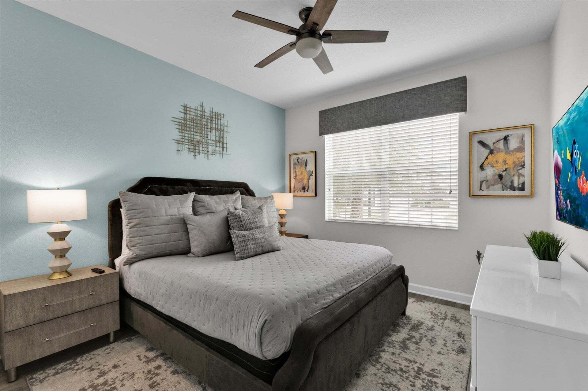 A bedroom with a bed, a ceiling fan, a nightstand, a bedside table, a window with blinds, and artwork on the walls.