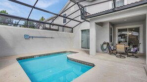 A swimming pool with a clear blue water, surrounded by a concrete deck. There is a covered patio area with a glass door leading to a room with a chair and a table. - Gas3070 (Kissimmee)
