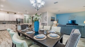 A modern kitchen with white cabinets, a dining area with a dark wooden table set for four, and a living area with a blue sofa and a TV. - Shd4742 (Kissimmee)