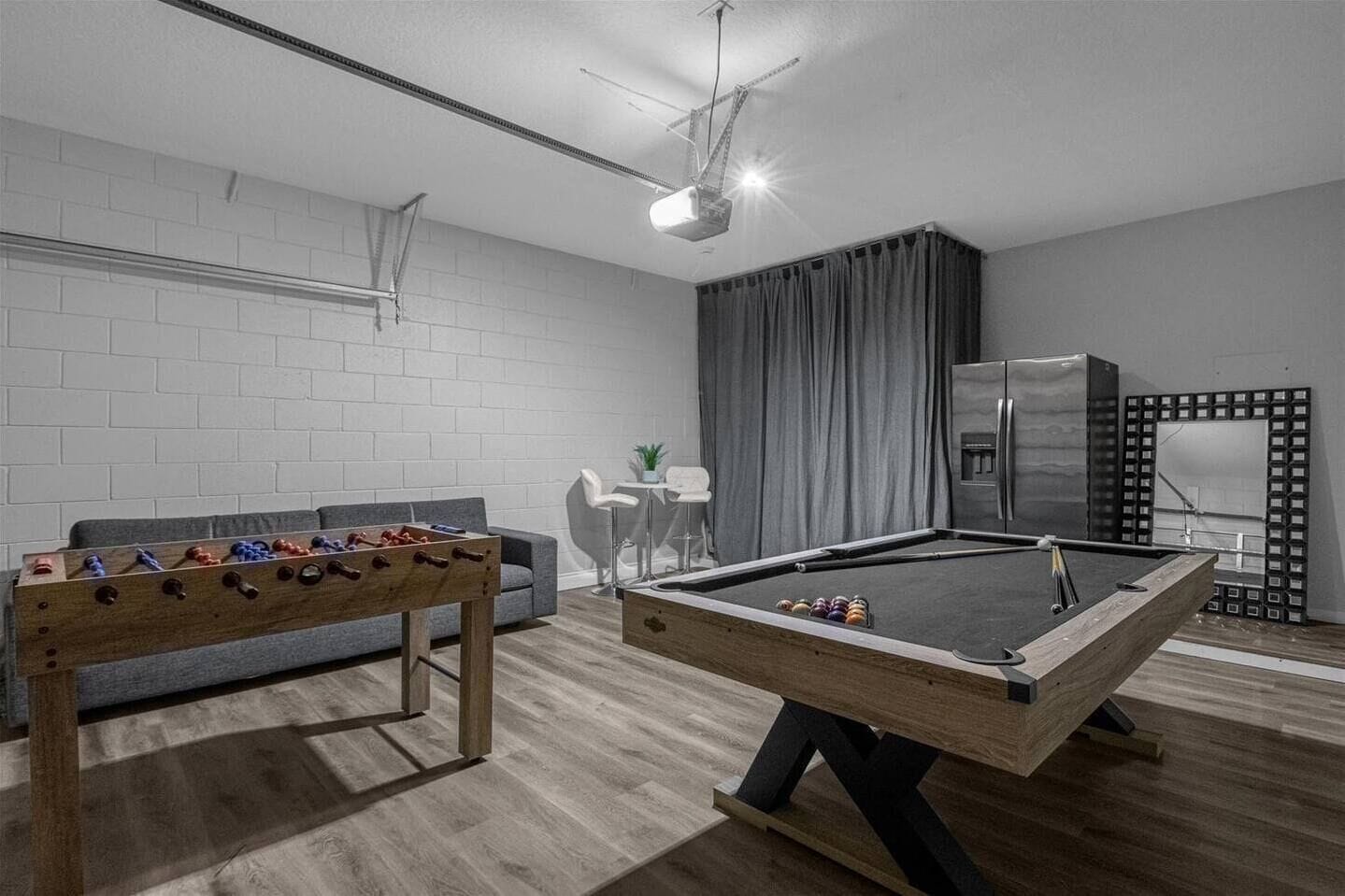 A modern living room with a foosball table, a pool table, a grey sofa, a small white table with a plant, and a refrigerator.