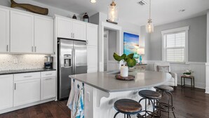 Una cocina moderna con armarios blancos, un refrigerador de acero inoxidable, una isla grande con taburetes, luces colgantes y vistas al área de estar con una ventana y un cuadro.