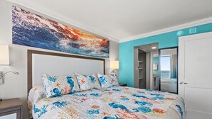 Quarto de hotel com uma cama e colcha com tema marinho, uma grande pintura abstrata acima da cama e vista para o mar por uma porta aberta.