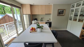 A modern kitchen with wooden cabinets, a white countertop, and a dining area with a table and chairs. - Arrowtown Central Apartment with Sundeck (Arrowtown)
