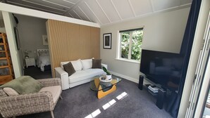 A living room with a white sofa, a glass coffee table, a TV, and a window with a view of greenery. - Arrowtown Central Apartment with Sundeck (Arrowtown)