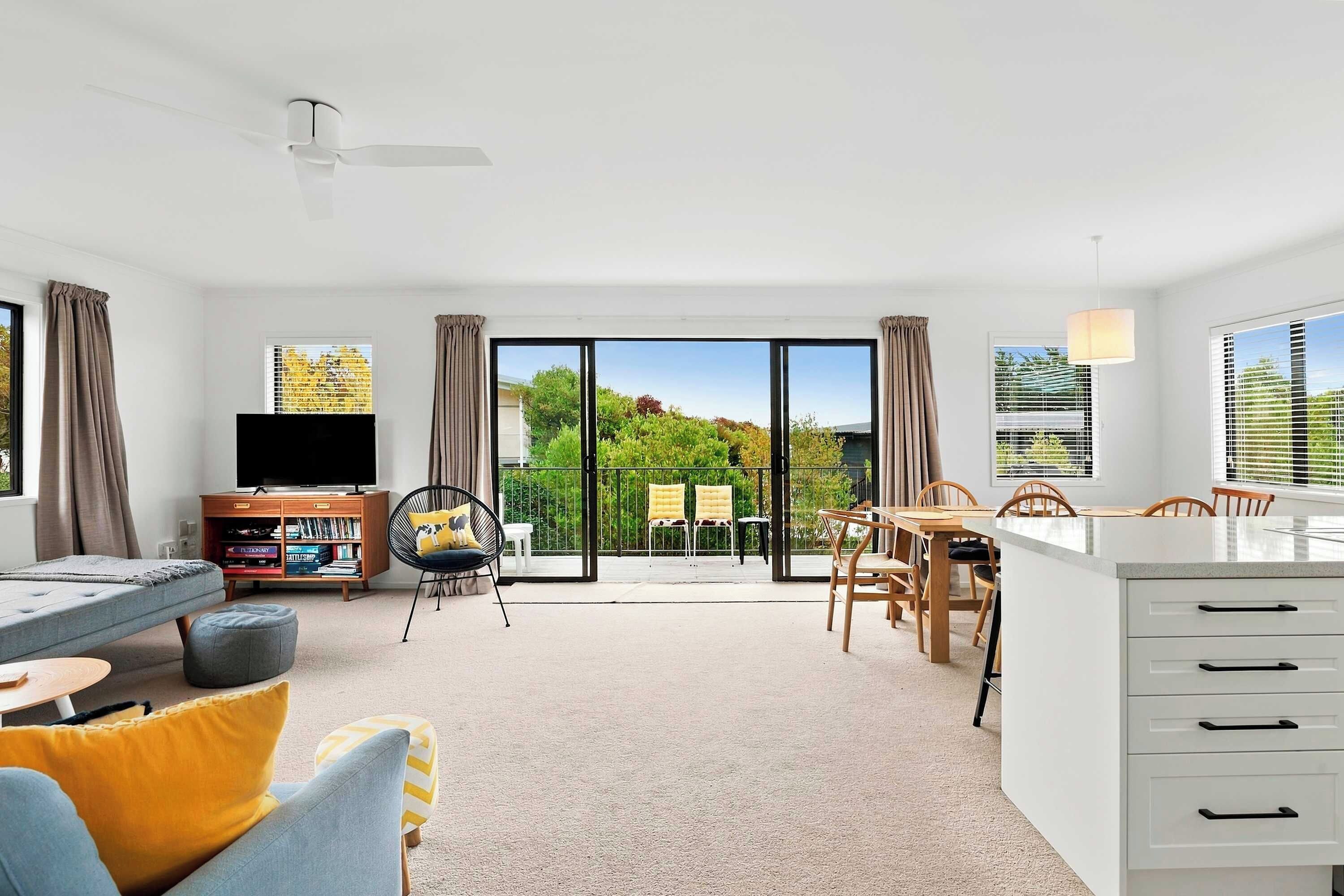A modern living room with a flat-screen TV, a dining area with a table and chairs, and a balcony with a view of greenery.